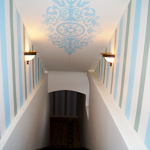Die Treppe zum Wellnessbereich führt vom historisierenden in den modernen Teil des Hauses. Das Deckenornament zitiert die klassische Formensprache, die Streifen der Wände sind modern.