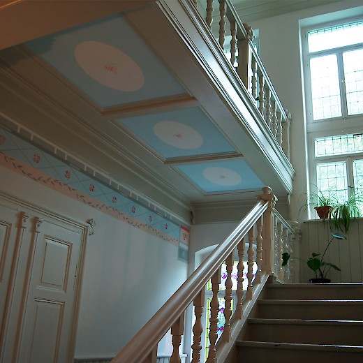 Auch die Deckenfelder der Treppe sind in blassblau. Wie eine Klammer zieht sich ein breites Ornamentband im oberen Wandbereich um den gesamten Raum, gibt ihm Halt und lenkt den Blick zur Decke